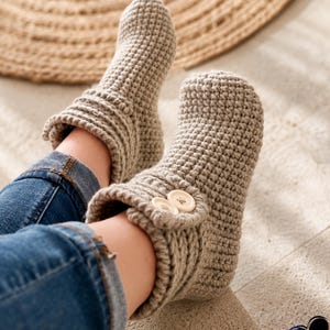 Könnte beinhalten: Ein Paar beige Häkelschuhe mit Knopfverschlüssen. Die Schuhe sind aus einem strukturierten Garn gefertigt und haben eine gerippte Manschette. Die Person trägt eine blaue Jeans. Ein runder gewebter Teppich befindet sich im Hintergrund.
