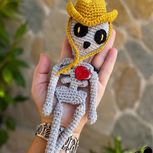 Puede incluir: Una figura de esqueleto tejida a ganchillo en hilo gris claro, con un sombrero de vaquero amarillo brillante, grandes cuencas oculares negras con pupilas amarillas y un pequeño corazón rojo cosido en el pecho. Cuerpo plano y articulado.