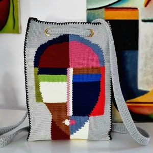 May include: A gray crochet shoulder bag with a colorful abstract face design. The face is composed of geometric shapes in shades of red, pink, blue, green, and white. The bag has a drawstring closure and a long strap.