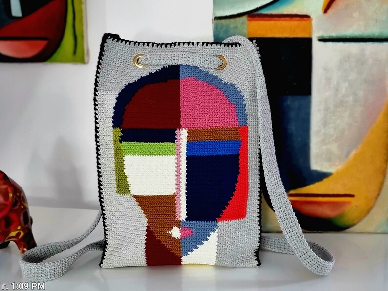 May include: A gray crochet shoulder bag with a colorful abstract face design. The face is composed of geometric shapes in shades of red, pink, blue, green, and white. The bag has a drawstring closure and a long strap.