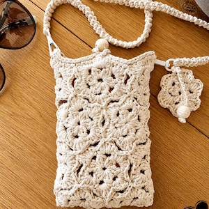 May include: A cream-colored, crocheted crossbody bag with a floral pattern. The bag has a long, braided strap and a small, matching charm. The bag is approximately 7 inches tall and 5 inches wide. Sunglasses are visible in the background.
