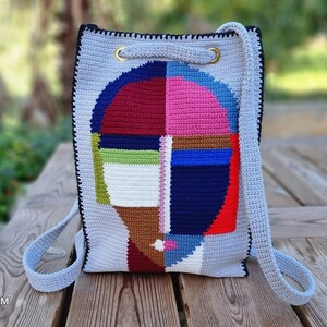 May include: A gray crochet backpack with a colorful abstract face design. The face is composed of geometric shapes in red, blue, pink, green, and brown. The bag has black trim, gold eyelets, and light blue straps.