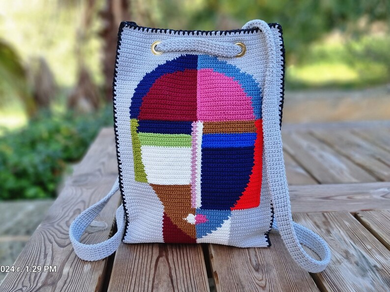 May include: A gray crochet backpack with a colorful abstract face design. The face is composed of geometric shapes in red, blue, pink, green, and brown. The bag has black trim, gold eyelets, and light blue straps.