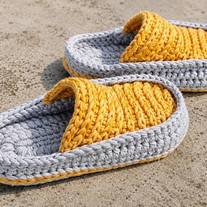 May include: A pair of handmade crochet slippers. The slippers feature a gray base with a yellow top and trim. The textured design is visible on the top of the slipper and the strap. The slippers are designed for indoor use.