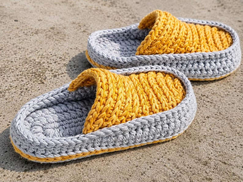 May include: A pair of handmade crochet slippers. The slippers feature a gray base with a yellow top and trim. The textured design is visible on the top of the slipper and the strap. The slippers are designed for indoor use.