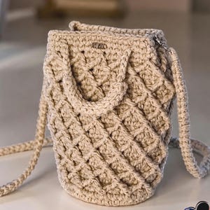 May include: A beige crochet handbag with a textured, diamond-patterned design. The bag has a cylindrical shape, a top handle, and a long, adjustable strap. A small metal tag is attached near the top. The bag is made of a soft, woven material.