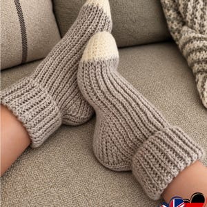 Könnte beinhalten: Ein Paar handgestrickte Socken mit gerippter Textur in Hellgrau. Die Socken verfügen über ein ausgeprägtes Bündchen am Knöchel und einen abgesetzten, abgerundeten Zehenbereich in Off-White. Das Strickmuster ist durchgehend gleichmäßig.