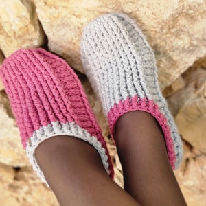 May include: A pair of handmade crochet slippers. One slipper is a vibrant pink, the other is a soft gray. Both have a pink trim around the opening and are made with a thick, textured yarn. The slippers are designed for indoor wear.