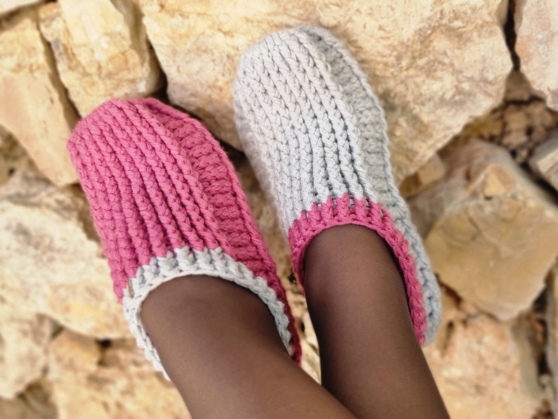 May include: A pair of handmade crochet slippers. One slipper is a vibrant pink, the other is a soft gray. Both have a pink trim around the opening and are made with a thick, textured yarn. The slippers are designed for indoor wear.