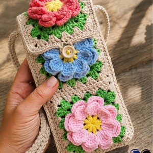 May include: A beige crochet phone bag adorned with three colorful crochet flowers. The flowers are pink, blue, and pink with yellow centers and green leaves. The bag has a button closure and a matching strap.
