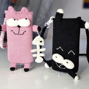 Op de afbeelding: Twee gehaakte telefoonhoesjes in de vorm van cartoonkatten. E&eacute;n is roze met witte en zwarte accenten, de andere is zwart met witte accenten. Beide hebben een riem en een kleine visgraatbedel. De roze kat heeft een lachend gezicht.