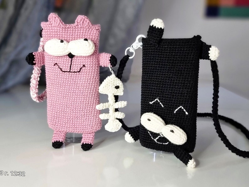 Op de afbeelding: Twee gehaakte telefoonhoesjes in de vorm van cartoonkatten. E&eacute;n is roze met witte en zwarte accenten, de andere is zwart met witte accenten. Beide hebben een riem en een kleine visgraatbedel. De roze kat heeft een lachend gezicht.