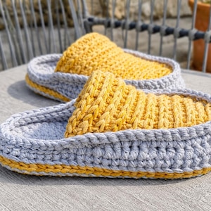 May include: A pair of handmade crochet slippers. The slippers feature a gray base with a yellow top and trim. The textured design is visible in the close-up shot, showcasing the craftsmanship. These cozy slippers are perfect for indoor wear.
