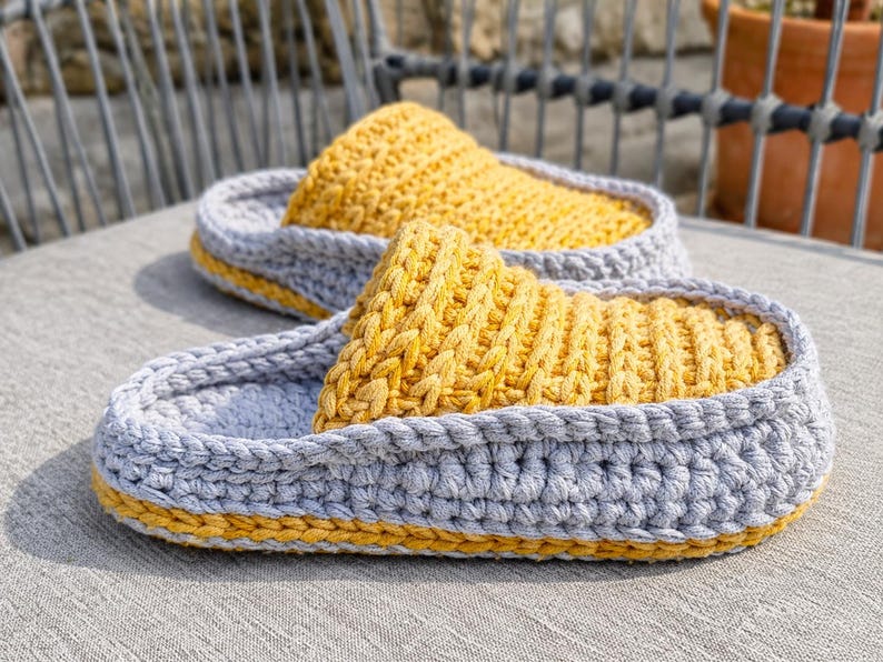 May include: A pair of handmade crochet slippers. The slippers feature a gray base with a yellow top and trim. The textured design is visible in the close-up shot, showcasing the craftsmanship. These cozy slippers are perfect for indoor wear.