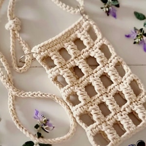 May include: Cream-colored macrame phone bag with a woven grid design. The bag has a long, braided strap and metal rings. The background includes purple flowers and green leaves, creating a natural aesthetic.