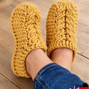 May include: A pair of mustard yellow slippers featuring a chunky knit texture with a vertical cable stitch pattern running down the center of each slipper. The slippers have a rounded toe design and a thick, textured yarn construction throughout.
