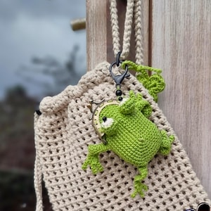 May include: A beige crochet drawstring bag with a green crochet frog attached. The frog has white eyes and is attached to a black clasp. The bag is hanging from a door handle.