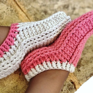 May include: A pair of handmade crochet socks in a soft, textured design. One sock is primarily light pink, the other is off-white. Both socks have a contrasting pink and off-white trim around the opening and toe area. The socks are shown on a person's feet.