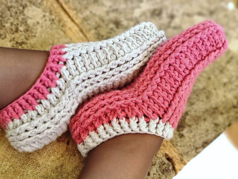 May include: A pair of handmade crochet socks in a soft, textured design. One sock is primarily light pink, the other is off-white. Both socks have a contrasting pink and off-white trim around the opening and toe area. The socks are shown on a person's feet.