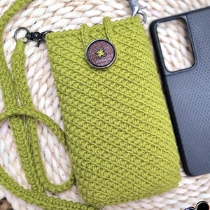 May include: A lime green crochet phone case with a matching strap and a brown button. The case is next to a black phone case on a woven surface. The case has a diagonal stitch pattern.