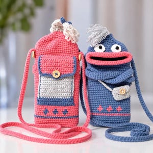 May include: Two crochet crossbody bags. The left bag is pink with a grey pocket, a wooden button, and a white tassel. The right bag is blue with a wide pink mouth, white eyes, grey hair, a blue scarf, and a small grey pouch with a wooden button.