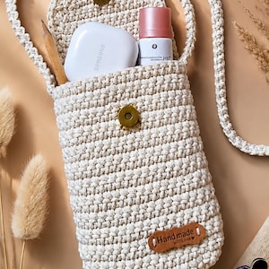 Könnte beinhalten: Eine cremefarbene, handgefertigte Häkeltasche mit Umhängegurt, Klappenverschluss und langem Riemen. Die Tasche enthält ein weißes Samsung-Etui, eine rosa-weiße Flasche und einen Bleistift. Ein kleines Lederetikett lautet "Hand made with love".