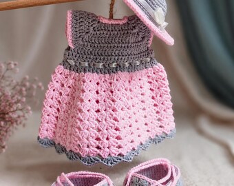 Puede incluir: Un conjunto tejido a mano en ganchillo que consta de un vestido gris y rosa con un sombrero gris a juego con cinta blanca, y un par de patucos grises y rosas con botones pequeños. El vestido tiene una falda rosa con patrón texturizado.