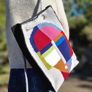 May include: A handmade, crocheted tote bag with a geometric design. The bag features a patchwork of colorful squares and shapes in red, pink, blue, green, and brown. The bag has a gray background, black trim, and a gray strap.