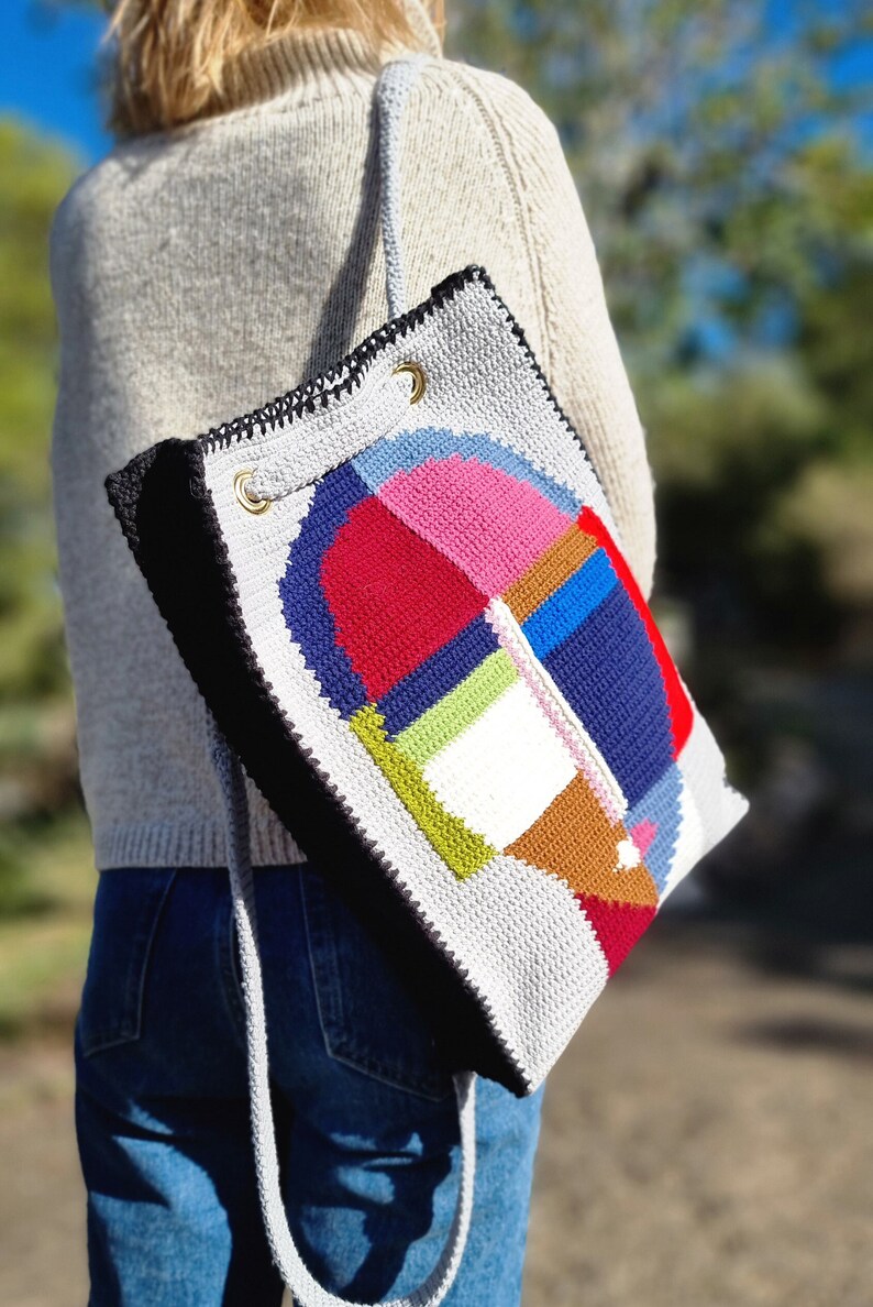 May include: A handmade, crocheted tote bag with a geometric design. The bag features a patchwork of colorful squares and shapes in red, pink, blue, green, and brown. The bag has a gray background, black trim, and a gray strap.
