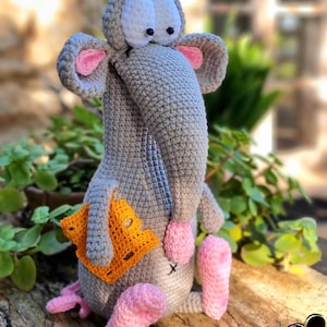Könnte beinhalten: Eine handgefertigte graue Häkelmauspuppe mit großen weißen Augen, rosa Ohren und rosa Füßen. Die Maus hält eine gelb-orangefarbene Käsescheibe aus Häkelarbeit. Die Puppe sitzt auf einer Holzoberfläche.