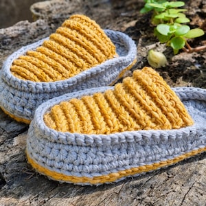 May include: A pair of handmade crochet slippers. The slippers feature a gray base with a yellow top and trim. The textured yellow top is ribbed, and the gray base has a woven appearance. The slippers are designed for indoor use.