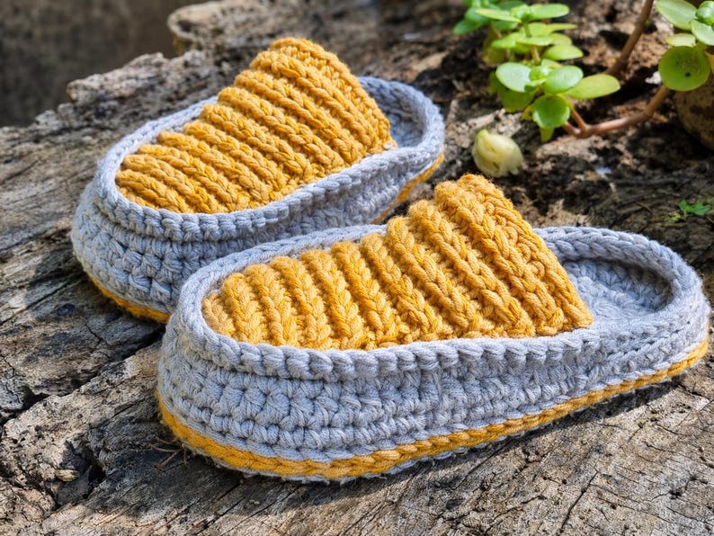 May include: A pair of handmade crochet slippers. The slippers feature a gray base with a yellow top and trim. The textured yellow top is ribbed, and the gray base has a woven appearance. The slippers are designed for indoor use.