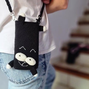 Op de afbeelding: Zwarte gehaakte telefoonhoes met een kattenontwerp. De hoes heeft een zwarte body met witte en cr&egrave;mekleurige accenten voor de ogen, poten en oren. Het heeft een zwarte band met zilverkleurige metalen sluitingen.