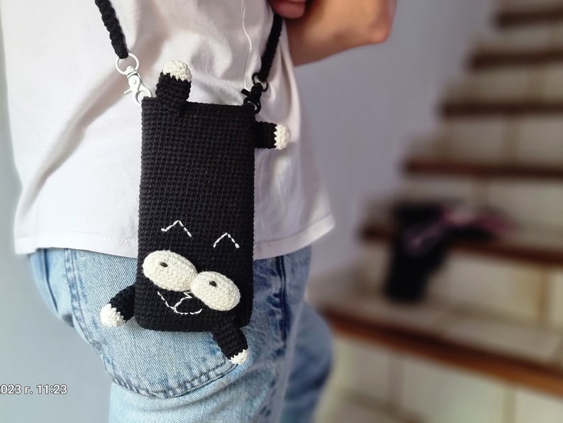 Op de afbeelding: Zwarte gehaakte telefoonhoes met een kattenontwerp. De hoes heeft een zwarte body met witte en cr&egrave;mekleurige accenten voor de ogen, poten en oren. Het heeft een zwarte band met zilverkleurige metalen sluitingen.