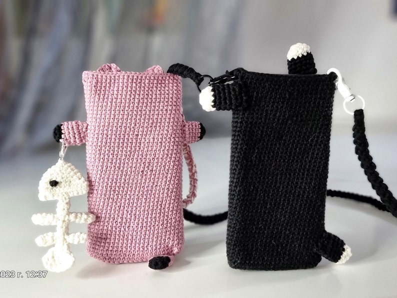 Op de afbeelding: Twee gehaakte telefoonhoesjes in de vorm van een kat. E&eacute;n is roze met zwarte accenten en een witte visgraatbedel. De andere is zwart met witte accenten en een zwarte koordriem. Beide hebben een rechthoekig ontwerp.