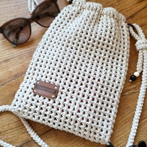Puede incluir: Un bolso de macramé hecho a mano de color crema, con cierre de cordón y una larga correa para el hombro. El bolso presenta una etiqueta rectangular de cuero que dice "HANDMADE". Un par de gafas de sol descansa junto al bolso. El bolso está diseñado con un patrón tejido.