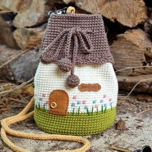 Könnte beinhalten: Eine handgefertigte, gehäkelte Tasche in Hausform mit braunem Dach, cremefarbenen Wänden und grünem Sockel. Die Tasche hat eine kleine braune Tür, einen Knopf und ein Blumendesign. Sie hat einen langen, gelben Riemen und ein Etikett mit der Aufschrift "Hand made with love".