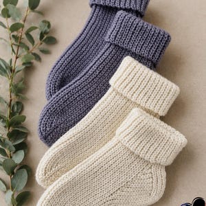 以下が含まれることがあります： 2組のニットソックス。1組はくすんだブルーグレー、もう1組はオフホワイトです。ソックスはリブ編みのカフスとテクスチャードニットパターンが特徴です。ニュートラルな背景に表示されています。