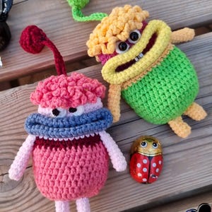Könnte beinhalten: Zwei handgefertigte Häkeltierpuppen in Rosa, Blau, Grün und Gelb. Eine Puppe hat einen rosafarbenen Körper, einen blauen Kragen und einen roten Stiel. Eine weitere Puppe ist grün mit gelben Haaren. Ein Marienkäfer-Schokoladen-Ei ist ebenfalls vorhanden.