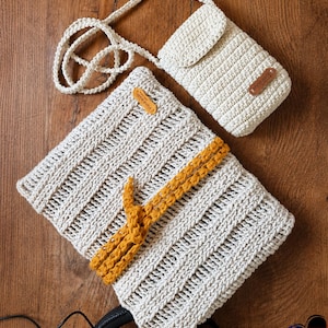 May include: Cream-colored crochet bag and matching crossbody phone case with a flap closure. The bag features a mustard yellow strap detail. Both items have small brown leather tags. The items are displayed on a wooden surface.