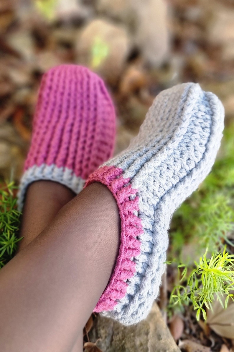 May include: A pair of handmade crochet slippers in a cozy design. One slipper is light gray with a pink trim, and the other is pink. The slippers are shown on a person's legs, with a blurred background of greenery.