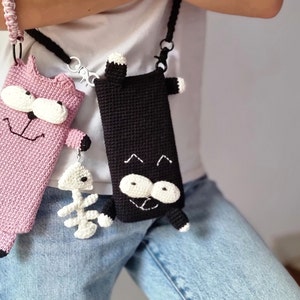 Op de afbeelding: Twee gehaakte telefoonhoesjes in de vorm van cartoonkatten. E&eacute;n is roze met zwarte accenten en een wit gezicht, de andere is zwart met witte accenten. Beide hebben bandjes en een visgraatbedel.