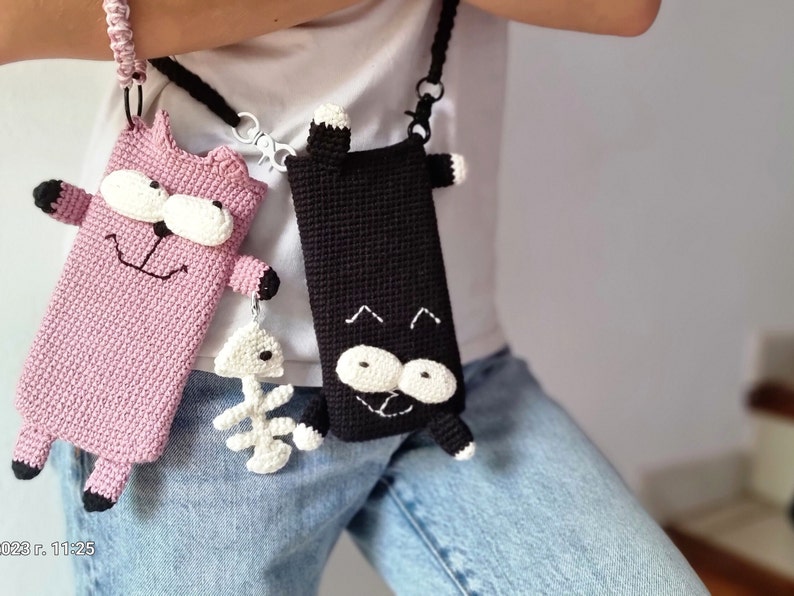 Op de afbeelding: Twee gehaakte telefoonhoesjes in de vorm van cartoonkatten. E&eacute;n is roze met zwarte accenten en een wit gezicht, de andere is zwart met witte accenten. Beide hebben bandjes en een visgraatbedel.