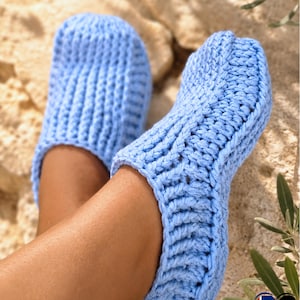 May include: A pair of light blue crocheted slippers. The slippers are made with a thick yarn and have a ribbed texture. The image shows the slippers on a person's feet, with the focus on the footwear. The slippers appear soft and comfortable.