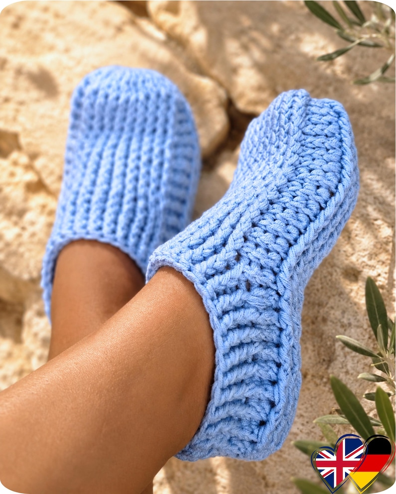 May include: A pair of light blue crocheted slippers. The slippers are made with a thick yarn and have a ribbed texture. The image shows the slippers on a person's feet, with the focus on the footwear. The slippers appear soft and comfortable.