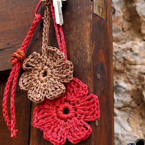 May include: A key hangs from a red braided lanyard, attached to a brown and red crocheted flower keychains. The keychains are in the shape of flowers. The background is a weathered wooden door and a stone wall.
