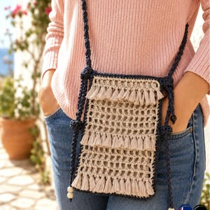 May include: A beige crochet crossbody bag with a navy blue strap and trim. The bag features a rectangular design with a grid pattern and three rows of beige fringe. The strap is adjustable and includes wooden beads.