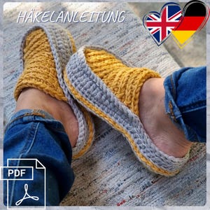 May include: Handmade yellow and grey crochet slippers. The slippers feature a thick, textured design with a grey border and yellow accents. The image also includes the text "HÄKELANLEITUNG" and a PDF icon.
