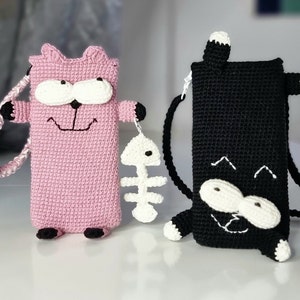 Op de afbeelding: Twee gehaakte telefoonhoesjes in de vorm van een kat. E&eacute;n is roze met een wit gezicht, zwarte accenten en een witte vissenbedel. De andere is zwart met een wit gezicht en accenten. Beide hebben riemen om te dragen.