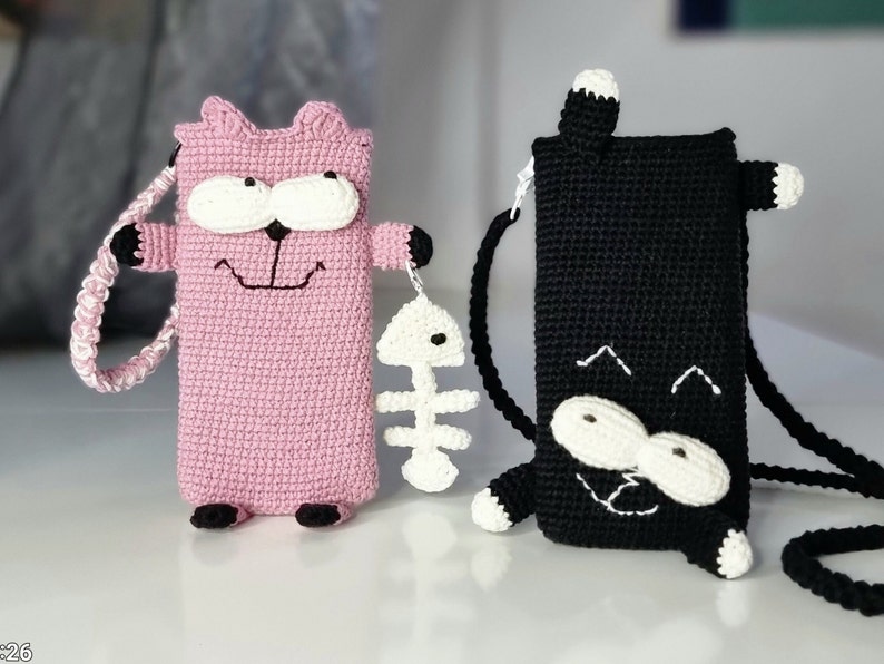 Op de afbeelding: Twee gehaakte telefoonhoesjes in de vorm van een kat. E&eacute;n is roze met een wit gezicht, zwarte accenten en een witte vissenbedel. De andere is zwart met een wit gezicht en accenten. Beide hebben riemen om te dragen.
